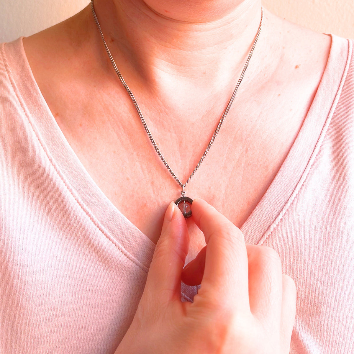 A close-up of a person wearing a delicate silver chain necklace with a small clear crystal pendant, touching the pendant with their fingers. The person is wearing a light pink V-neck top against a soft beige background.