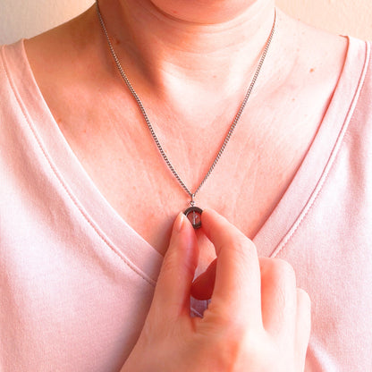 A close-up of a person wearing a delicate silver chain necklace with a small clear crystal pendant, touching the pendant with their fingers. The person is wearing a light pink V-neck top against a soft beige background.