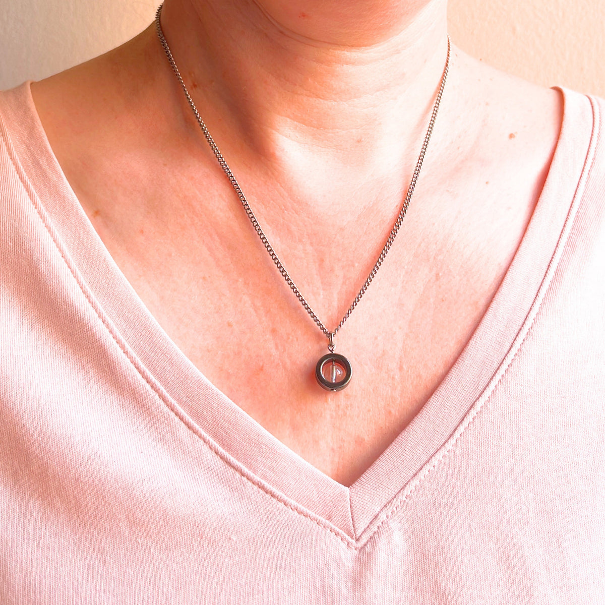 A close-up of a person's neck and chest wearing a delicate silver chain necklace with a small circular pendant featuring a clear crystal gemstone, paired with a light pink V-neck top.