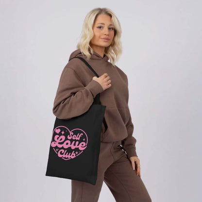 Woman wearing a brown hoodie and pants holding a black tote bag with 'Self Love Club' text.