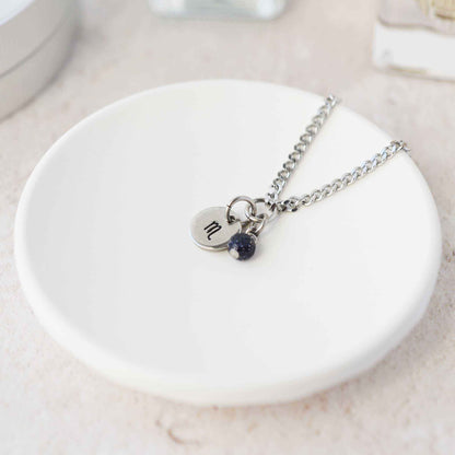 Tiny Scorpio zodiac sign disc pendant necklace with Blue Goldstone charm laying on white trinket dish
