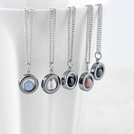 Five different coloured dainty gemstone spinner necklaces hanging on white cup.