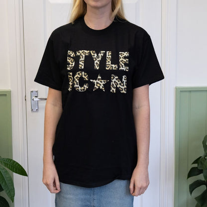 Person wearing a black t-shirt with 'STYLE ICON' text in leopard print, standing in front of a white door.