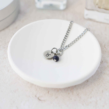 Tiny Taurus zodiac sign disc pendant necklace with Blue Goldstone charm laying on white trinket dish