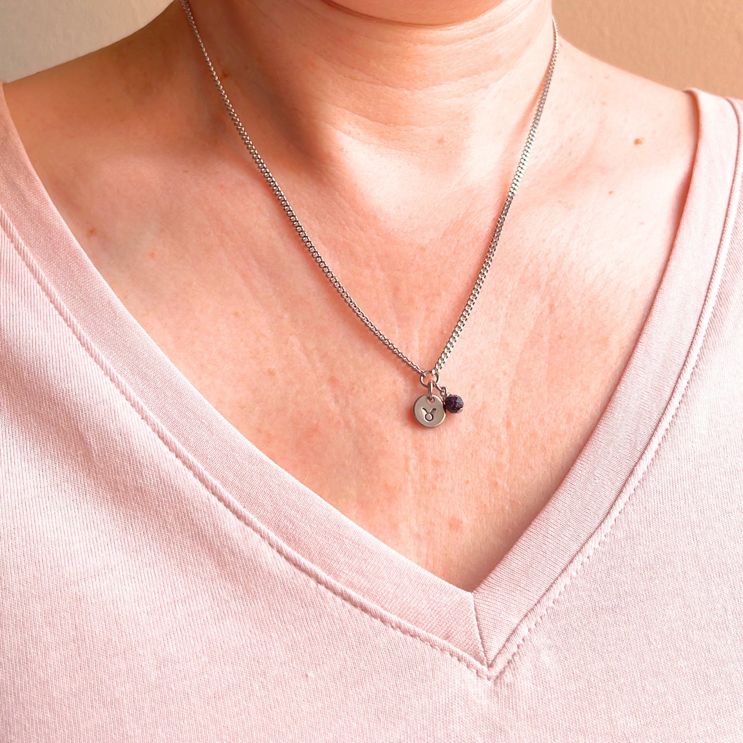 Woman wearing a pink v neck top and dainty Taurus zodiac sign necklace.