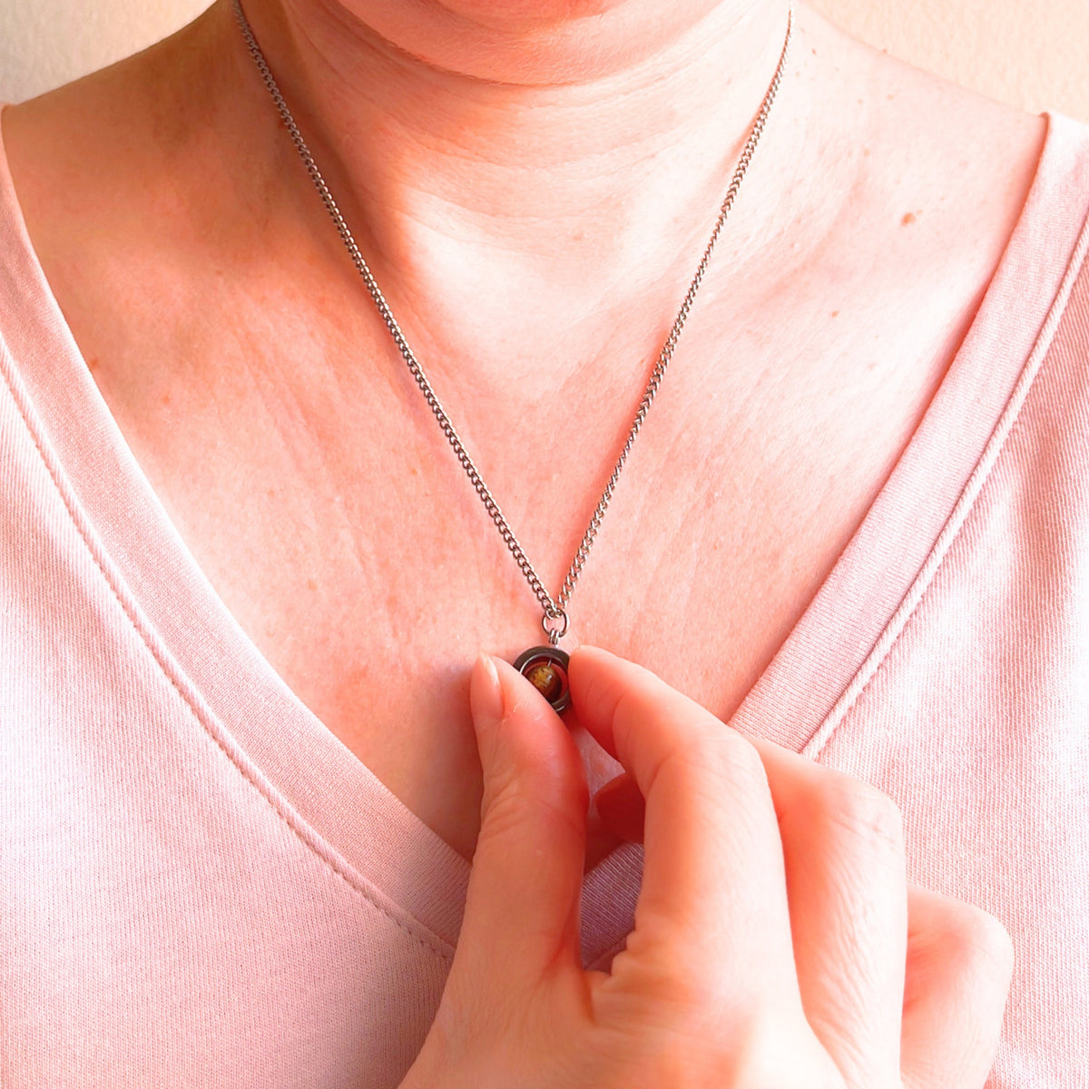 A close-up of a person wearing a delicate silver chain necklace with a small gemstone pendant, while a hand gently touches the pendant. The person is wearing a light pink V-neck top.