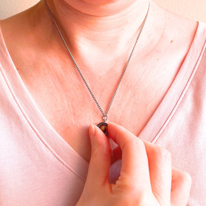 A close-up of a person wearing a delicate silver chain necklace with a small gemstone pendant, while a hand gently touches the pendant. The person is wearing a light pink V-neck top.