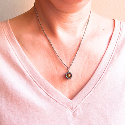 A close-up of a person's neck and chest wearing a silver chain necklace with a circular pendant featuring a Tiger's Eye gemstone, against a light pink V-neck top.