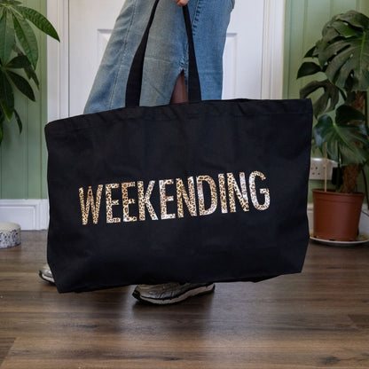 Black tote bag with 'WEEKENDING' text held by a person indoors.