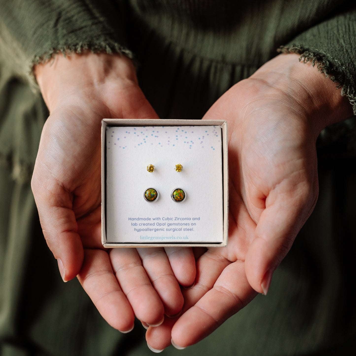 Hands holding a square gift box with small and large yellow green stud earrings inside.
