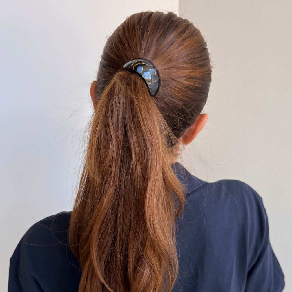 Person with long brown hair tied in a ponytail wearing a dark blue shirt against a light background