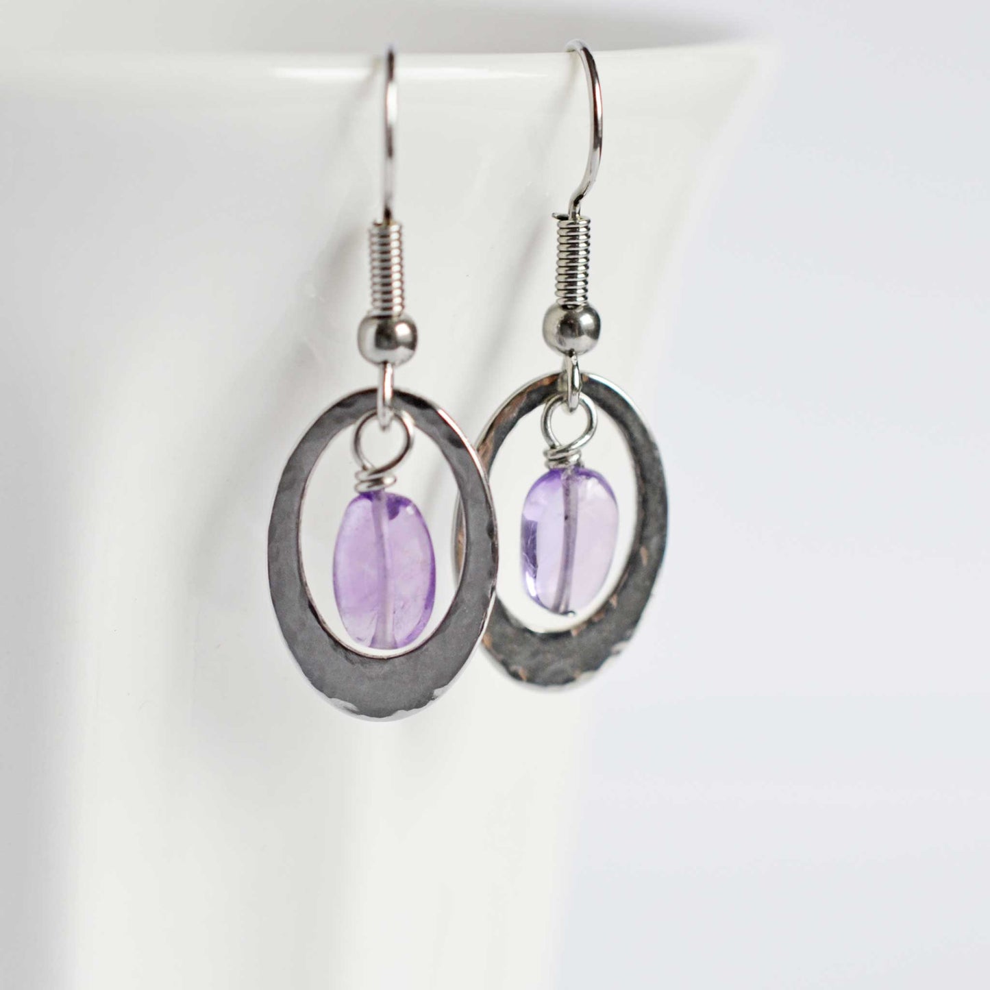 Hammered oval drop earrings with central Amethyst gemstone hanging on white cup.