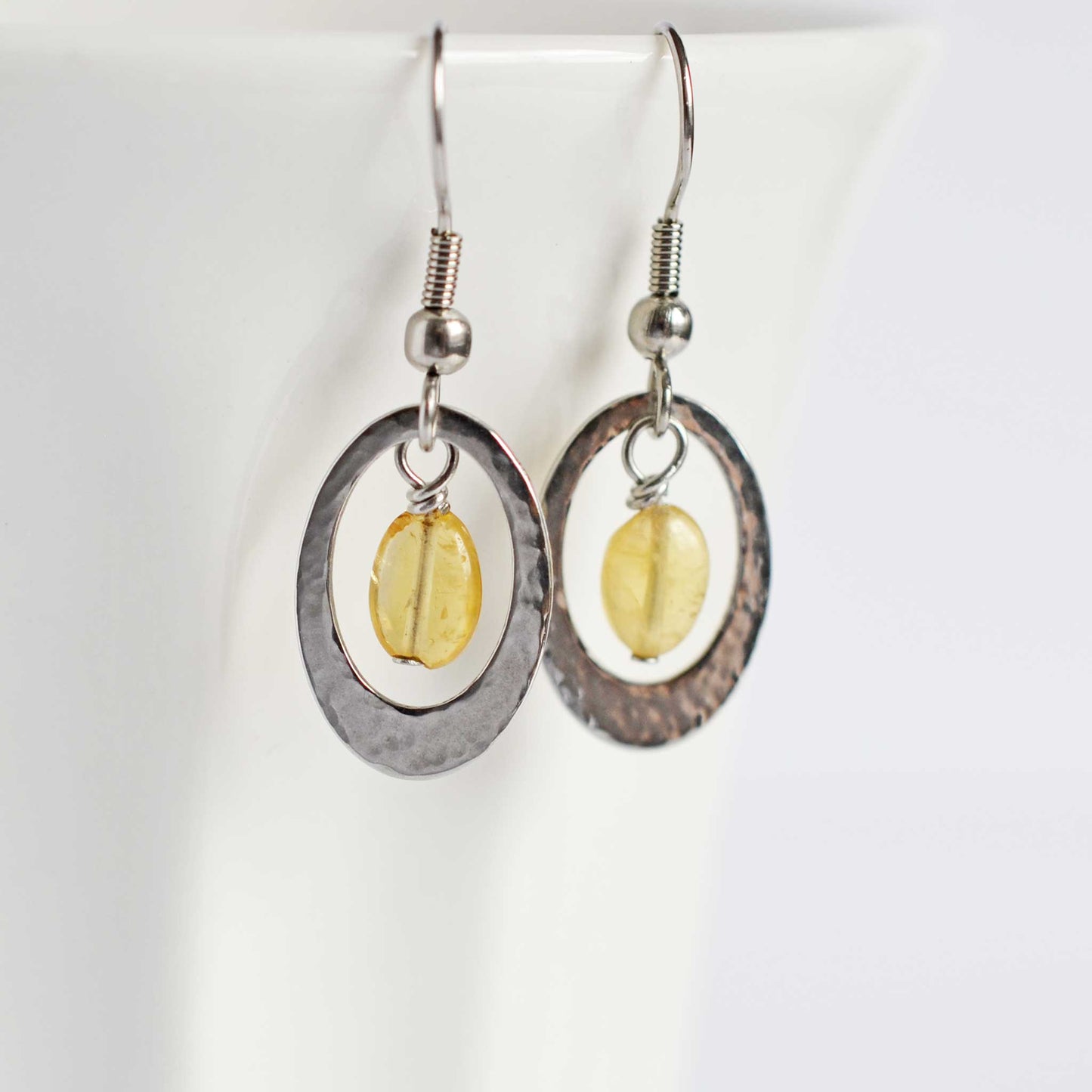 Pair of steel oval and yellow Citrine earrings hanging on white cup.