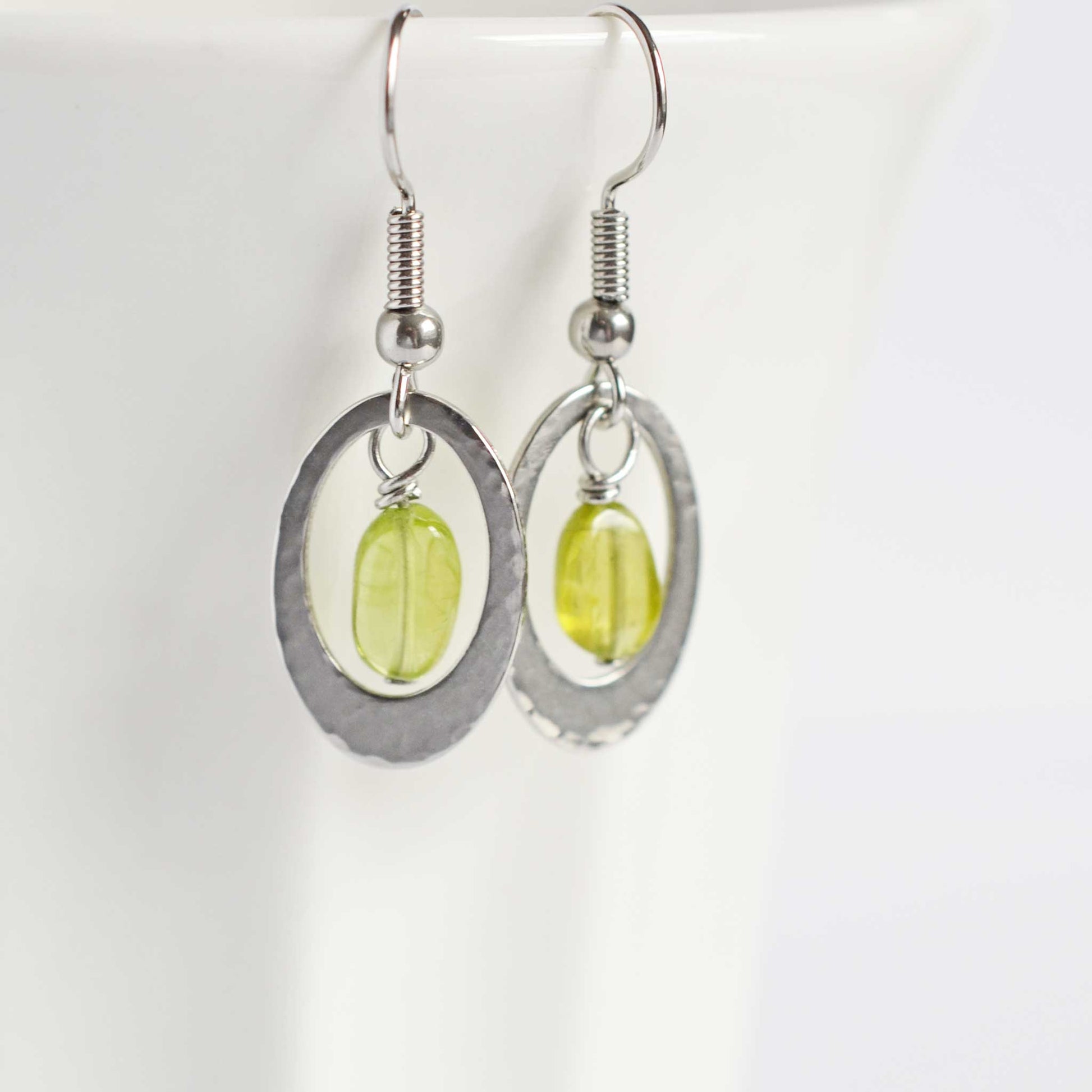 Green Peridot and steel oval drop earrings hanging on white cup.